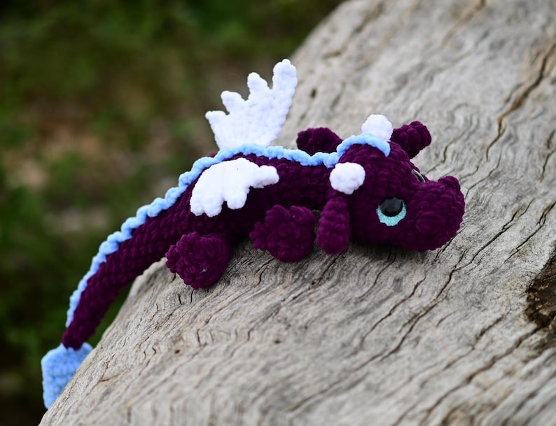 May include: A crocheted dragon plush toy featuring a deep purple body, white wings, and a light blue spine ridge. The creature has large, dark eyes with light blue irises, small horns on its head, and a long tail that ends in a light blue tip.