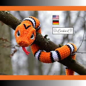 Puede incluir: Un juguete de serpiente tejido a ganchillo con un patrón de rayas naranjas, negras y blancas. La serpiente de peluche tiene ojos grandes y brillantes, una lengua de hilo rojo y una marca de chevron negra en la cabeza. Se ve el texto Crochimal.