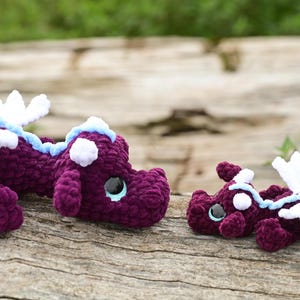 May include: Two crocheted dragon plushies made of dark purple yarn with white wings, light blue accents along the back, and large black safety eyes. The larger dragon is positioned on the left, while the smaller dragon is on the right.