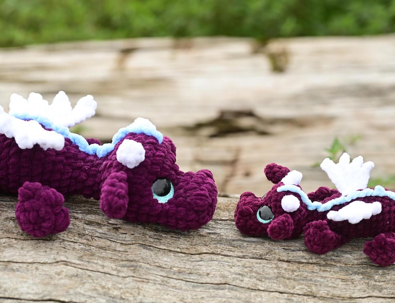 May include: Two crocheted dragon plushies made of dark purple yarn with white wings, light blue accents along the back, and large black safety eyes. The larger dragon is positioned on the left, while the smaller dragon is on the right.
