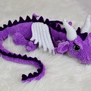 K&ouml;nnte beinhalten: Ein geh&auml;keltes lila Drachenspielzeug mit wei&szlig;en Fl&uuml;geln und H&ouml;rnern. Der Drache liegt auf einer wei&szlig;en, flauschigen Oberfl&auml;che. Der K&ouml;rper des Drachens ist ein leuchtendes Lila mit dunkleren lila Akzenten entlang des R&uuml;ckens und des Schwanzes.