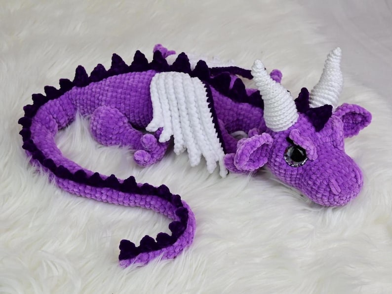 K&ouml;nnte beinhalten: Ein geh&auml;keltes lila Drachenspielzeug mit wei&szlig;en Fl&uuml;geln und H&ouml;rnern. Der Drache liegt auf einer wei&szlig;en, flauschigen Oberfl&auml;che. Der K&ouml;rper des Drachens ist ein leuchtendes Lila mit dunkleren lila Akzenten entlang des R&uuml;ckens und des Schwanzes.