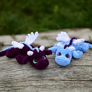 May include: Two handmade crochet dragon plushies. One dragon is deep purple with light blue accents and white wings, while the other is light blue with a dark purple spine and white wings. Both feature large dark eyes and soft yarn textures.
