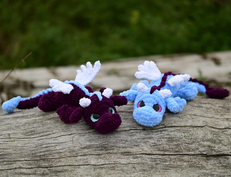 May include: Two handmade crochet dragon plushies. One dragon is deep purple with light blue accents and white wings, while the other is light blue with a dark purple spine and white wings. Both feature large dark eyes and soft yarn textures.