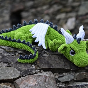 K&ouml;nnte beinhalten: Ein handgefertigter gr&uuml;ner H&auml;keldrachen mit wei&szlig;en Fl&uuml;geln, H&ouml;rnern und dunkelblauen Akzenten. Der Drache liegt auf einer Steinoberfl&auml;che. Der Schwanz und der R&uuml;cken des Drachen sind mit dunkelblauen Stacheln versehen.