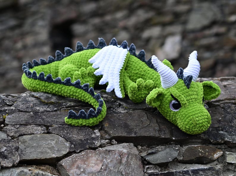 K&ouml;nnte beinhalten: Ein handgefertigter gr&uuml;ner H&auml;keldrachen mit wei&szlig;en Fl&uuml;geln, H&ouml;rnern und dunkelblauen Akzenten. Der Drache liegt auf einer Steinoberfl&auml;che. Der Schwanz und der R&uuml;cken des Drachen sind mit dunkelblauen Stacheln versehen.