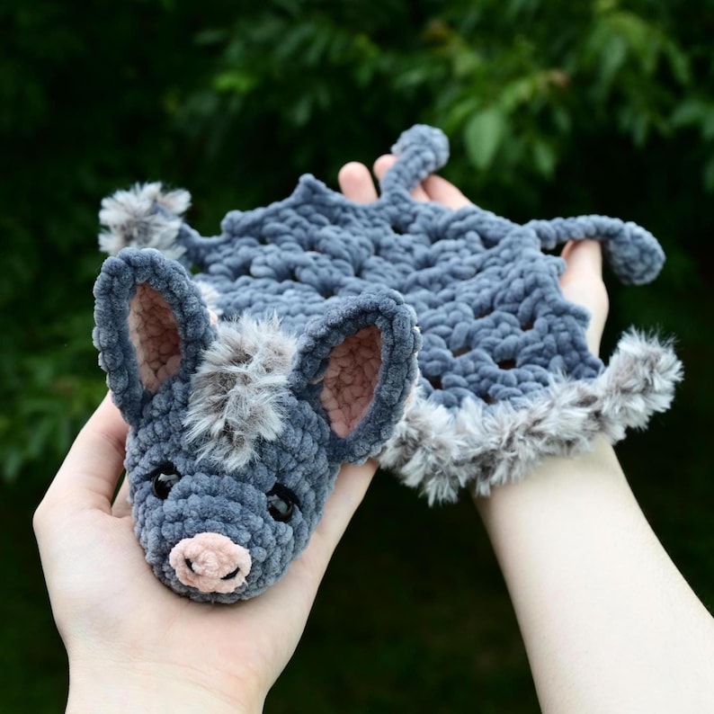 May include: A handmade crochet bat-shaped baby blanket in shades of gray. The bat's head features large ears with pink inner ears, a fluffy gray snout, and small black eyes. The blanket has a textured, plush appearance with a faux fur trim.