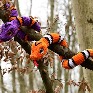 Pode incluir: Dois brinquedos de cobra de croch&ecirc;, um roxo e laranja, o outro laranja e branco, est&atilde;o enrolados em torno de um galho de &aacute;rvore.