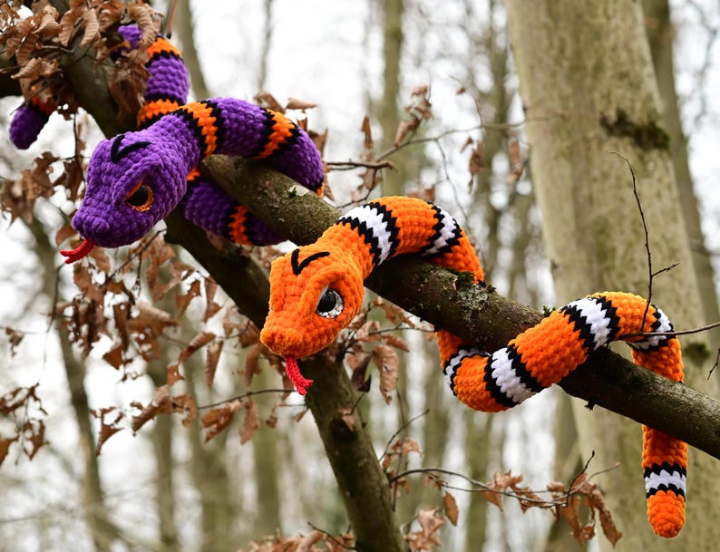 Pode incluir: Dois brinquedos de cobra de croch&ecirc;, um roxo e laranja, o outro laranja e branco, est&atilde;o enrolados em torno de um galho de &aacute;rvore.