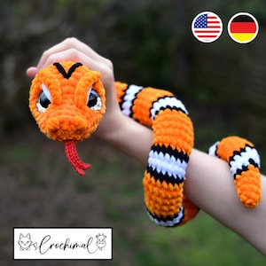 Pode incluir: Um brinquedo de cobra de croch&ecirc; laranja e preto com listras brancas. A cobra tem uma l&iacute;ngua vermelha e olhos pretos. A cobra est&aacute; sendo segurada pelo bra&ccedil;o de uma pessoa.