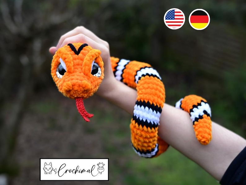 Pode incluir: Um brinquedo de cobra de croch&ecirc; laranja e preto com listras brancas. A cobra tem uma l&iacute;ngua vermelha e olhos pretos. A cobra est&aacute; sendo segurada pelo bra&ccedil;o de uma pessoa.