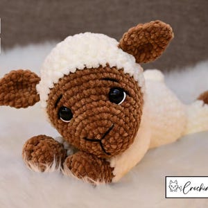 May include: A brown and white crocheted lamb toy with black eyes. The lamb is lying on a white fluffy surface.