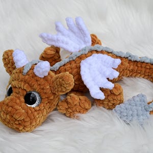 May include: A plush dragon toy crafted from soft brown yarn, featuring white wings, white horns, and a light gray spine and tail tip. The creature has large, round, black safety eyes with silver glitter accents, giving it a distinct appearance.