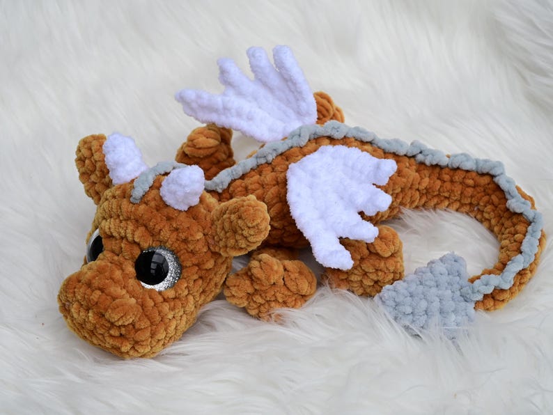 May include: A plush dragon toy crafted from soft brown yarn, featuring white wings, white horns, and a light gray spine and tail tip. The creature has large, round, black safety eyes with silver glitter accents, giving it a distinct appearance.