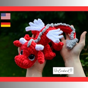 May include: A handmade crochet dragon plush toy featuring a vibrant red body, white wings, and white horns. The creature has large black eyes with silver glitter accents and a grey spine and tail tip. A small white label displays the text Crochimal.