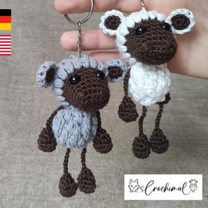 Puede incluir: Dos llaveros de oveja tejidos a crochet, uno gris y otro blanco, con caras y patas marrones. Los llaveros cuelgan de un llavero plateado.