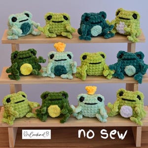 Op de afbeelding: Een verzameling van 10 gehaakte kikker knuffels in verschillende tinten groen, blauw en geel. De kikkers zijn gerangschikt op een houten plank met de tekst "no sew" op de onderste plank.