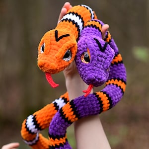 Pode incluir: Duas cobras de croch&ecirc;, uma laranja e branca, a outra roxa e preta, est&atilde;o entrela&ccedil;adas. As cobras t&ecirc;m olhos pretos e l&iacute;nguas vermelhas.