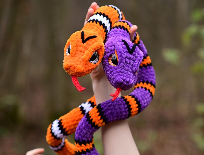 Pode incluir: Duas cobras de croch&ecirc;, uma laranja e branca, a outra roxa e preta, est&atilde;o entrela&ccedil;adas. As cobras t&ecirc;m olhos pretos e l&iacute;nguas vermelhas.