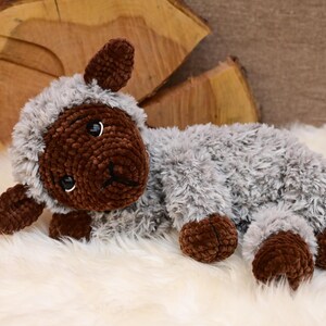 May include: A crocheted plush toy lamb with brown fur on the face, legs, and ears, and grey fur on the body. The lamb is lying on a white fluffy surface.