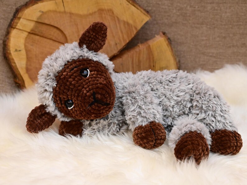 May include: A crocheted plush toy lamb with brown fur on the face, legs, and ears, and grey fur on the body. The lamb is lying on a white fluffy surface.