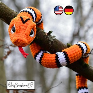 Pode incluir: Uma cobra de croch&ecirc; laranja e preta com listras brancas, com olhos grandes e uma l&iacute;ngua vermelha, est&aacute; enrolada em volta de um galho de &aacute;rvore. A cobra &eacute; um brinquedo de pel&uacute;cia feito &agrave; m&atilde;o.
