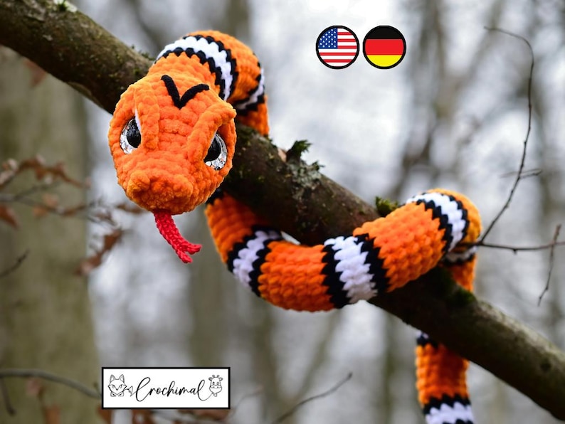 Pode incluir: Uma cobra de croch&ecirc; laranja e preta com listras brancas, com olhos grandes e uma l&iacute;ngua vermelha, est&aacute; enrolada em volta de um galho de &aacute;rvore. A cobra &eacute; um brinquedo de pel&uacute;cia feito &agrave; m&atilde;o.