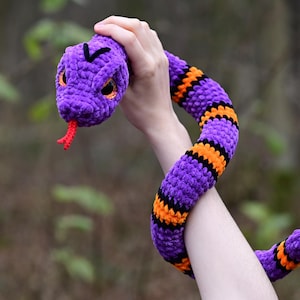 Pode incluir: Um brinquedo de cobra de croch&ecirc; roxo e laranja com uma l&iacute;ngua vermelha. A cobra est&aacute; sendo segurada pela m&atilde;o de uma pessoa.