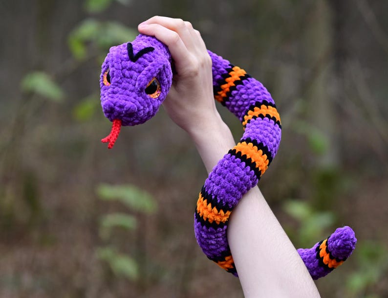Pode incluir: Um brinquedo de cobra de croch&ecirc; roxo e laranja com uma l&iacute;ngua vermelha. A cobra est&aacute; sendo segurada pela m&atilde;o de uma pessoa.