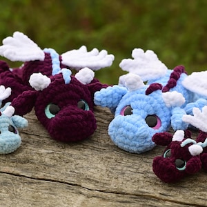 May include: A collection of four crocheted dragon plush toys in varying sizes. Two dragons are deep purple with light blue accents, and two are light blue with dark purple accents. Each features white wings, small horns, and large black eyes.