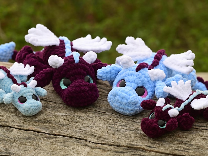 May include: A collection of four crocheted dragon plush toys in varying sizes. Two dragons are deep purple with light blue accents, and two are light blue with dark purple accents. Each features white wings, small horns, and large black eyes.