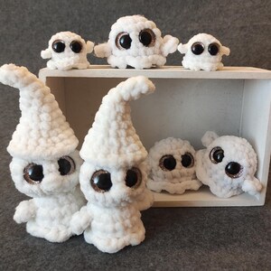 May include: A group of white crocheted ghost plushies with large googly eyes. The ghosts are arranged in a wooden box with a white interior.