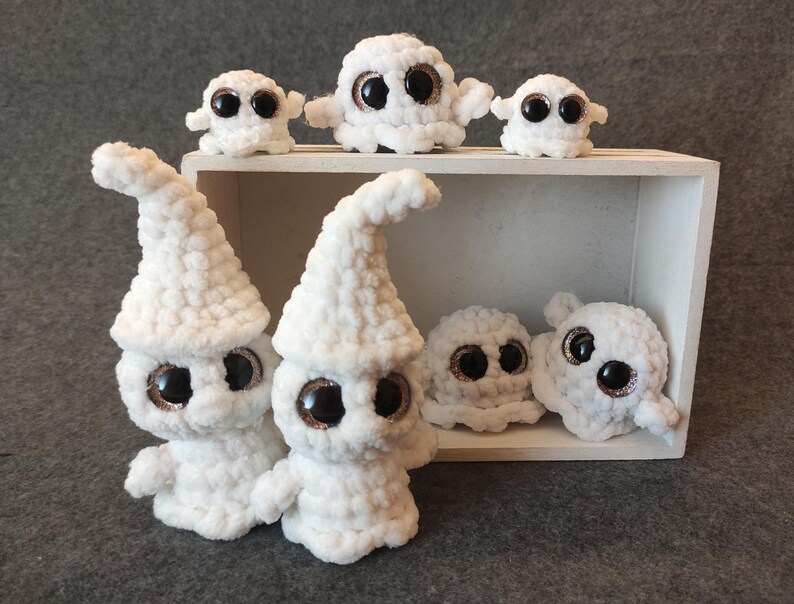 May include: A group of white crocheted ghost plushies with large googly eyes. The ghosts are arranged in a wooden box with a white interior.