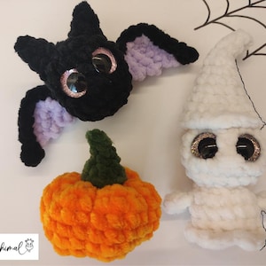 Könnte beinhalten: Drei gehäkelte Halloween-Dekorationen: eine schwarze Fledermaus mit lila Flügeln, ein weißer Geist mit Glitz Augen und ein orangefarbener Kürbis mit grünem Stiel.