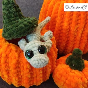 May include: A crocheted gray and white dragon with large eyes sits inside a crocheted orange pumpkin. The pumpkin is decorated with a green crocheted hat. The image includes the text "Crochimal".