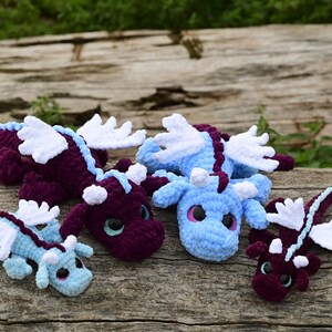 May include: Four crocheted dragon plush toys in varying sizes and colors. Two are deep purple with light blue accents, and two are light blue with purple accents. Each features white wings, small horns, and large, dark, glossy eyes.