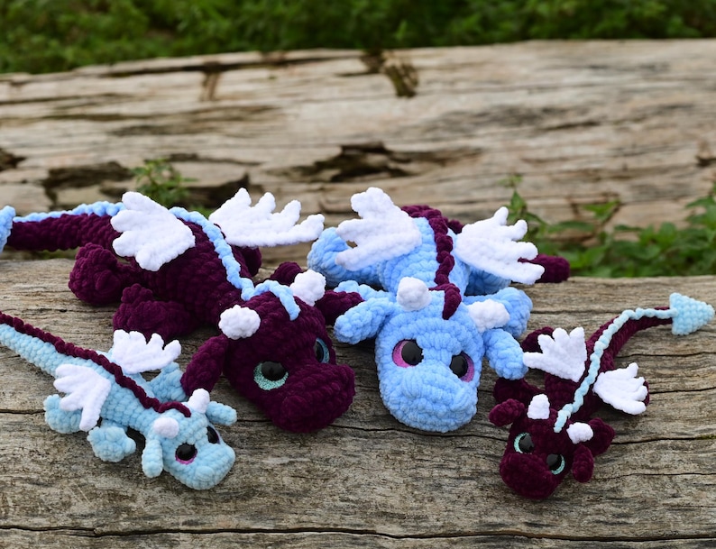May include: Four crocheted dragon plush toys in varying sizes and colors. Two are deep purple with light blue accents, and two are light blue with purple accents. Each features white wings, small horns, and large, dark, glossy eyes.