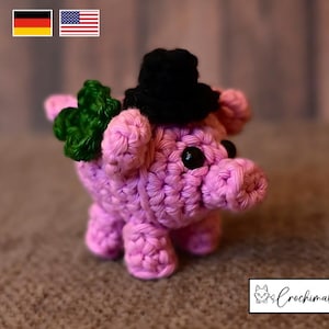 Könnte beinhalten: Ein handgehäkeltes rosa Ferkel mit schwarzem Zylinder und grünem Kleeblatt. Das Ferkel hat schwarze Knopfaugen und ist ein skurriles, handgefertigtes Objekt. Das Bild enthält die deutsche und die amerikanische Flagge.