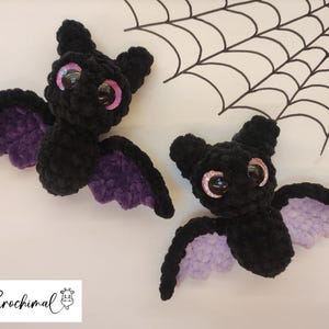Pode incluir: Dois morcegos pretos de crochê com asas roxas e olhos grandes e brilhantes. Os morcegos estão posicionados em uma superfície branca com um desenho de teia de aranha no fundo. Os morcegos são feitos à mão.