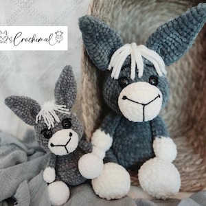 May include: Two crocheted gray donkey plushies with white faces and white yarn for manes. The larger donkey is sitting on a woven basket, while the smaller donkey is sitting on a gray blanket.