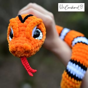 Pode incluir: Uma cobra de croch&ecirc; laranja e preta com listras brancas e grandes olhos saltados. A cobra tem uma l&iacute;ngua vermelha.