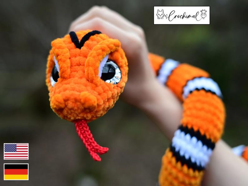 Pode incluir: Uma cobra de croch&ecirc; laranja e preta com listras brancas e grandes olhos saltados. A cobra tem uma l&iacute;ngua vermelha.