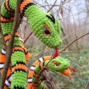Pode incluir: Duas cobras de croch&ecirc; verde com listras laranja e pretas. As cobras t&ecirc;m l&iacute;nguas vermelhas e olhos grandes e pretos. As cobras est&atilde;o enroladas em torno de um galho.