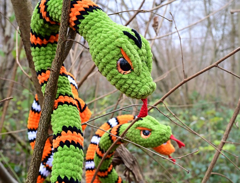 Pode incluir: Duas cobras de croch&ecirc; verde com listras laranja e pretas. As cobras t&ecirc;m l&iacute;nguas vermelhas e olhos grandes e pretos. As cobras est&atilde;o enroladas em torno de um galho.