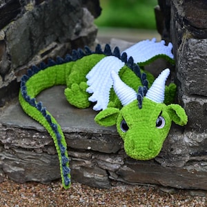 K&ouml;nnte beinhalten: Ein handgefertigtes gr&uuml;nes H&auml;keldrachen-Spielzeug mit wei&szlig;en Fl&uuml;geln und H&ouml;rnern. Der Drache hat einen langen Schwanz mit dunkelblauen Akzenten und ruht auf einer Steinoberfl&auml;che. Die Augen des Drachens sind dunkel und er hat einen leicht m&uuml;rrischen Ausdruck.