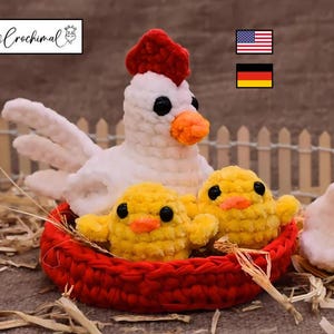 May include: A handmade crochet hen with a red comb, white body, and orange beak, stands over a red basket holding two yellow chicks. The scene is set on a bed of straw, with a small fence in the background.