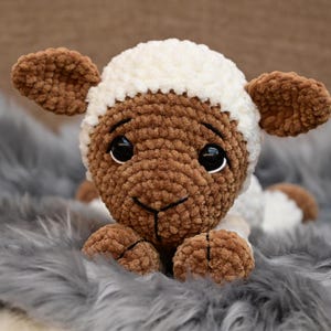 May include: A brown and white crocheted lamb plush toy with black eyes. The lamb is lying on a grey fluffy surface.