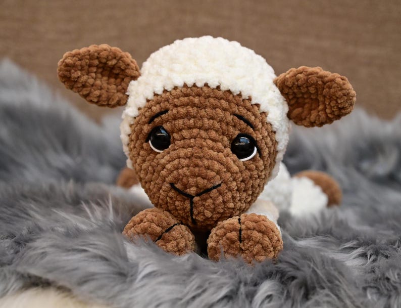 May include: A brown and white crocheted lamb plush toy with black eyes. The lamb is lying on a grey fluffy surface.