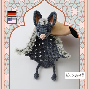 May include: A handmade crochet bat plushie in shades of gray and black, with a pink nose and inner ears. The bat has large ears and outstretched wings. The plushie is held against a patterned background. The logo "Crochimal" is visible.