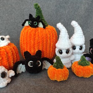 May include: A collection of crocheted Halloween decorations, including pumpkins, ghosts, and bats. The pumpkins are orange and have a fuzzy texture. The ghosts are white and have googly eyes. The bats are black with purple wings.